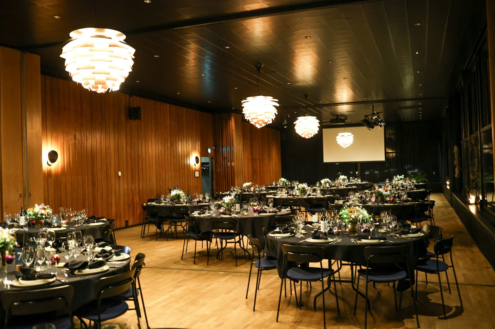 Corporate gala dinner with round tables set beneath pendant lights and a presentation screen