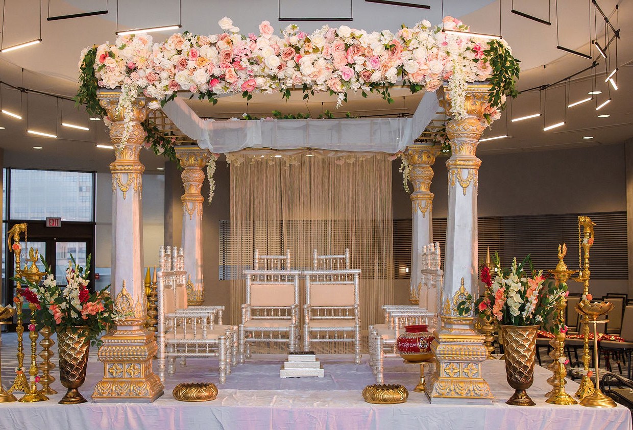 Wedding mandap with a pink and white floral canopy, carved pillars and gold lamps