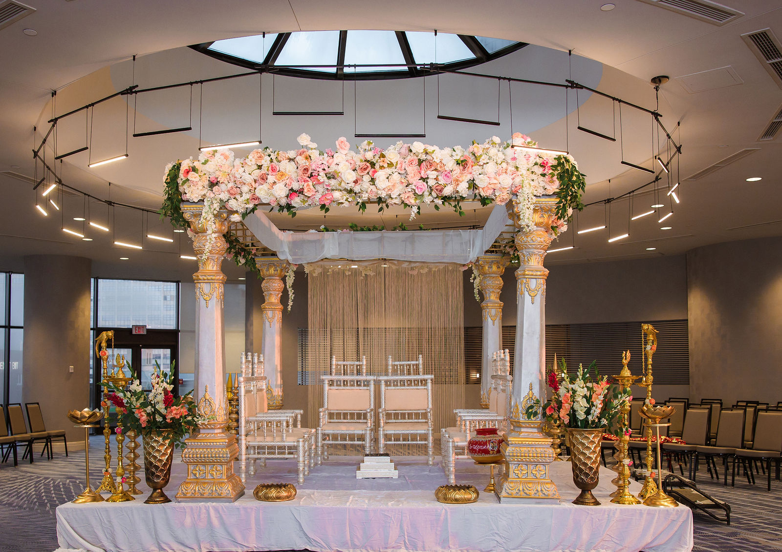Mandap with a pink and white floral canopy, carved pillars and gold urns under a skylight