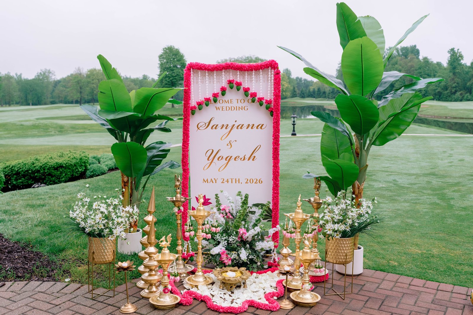 Floral welcome sign with brass lamps and palms on a golf course lawn