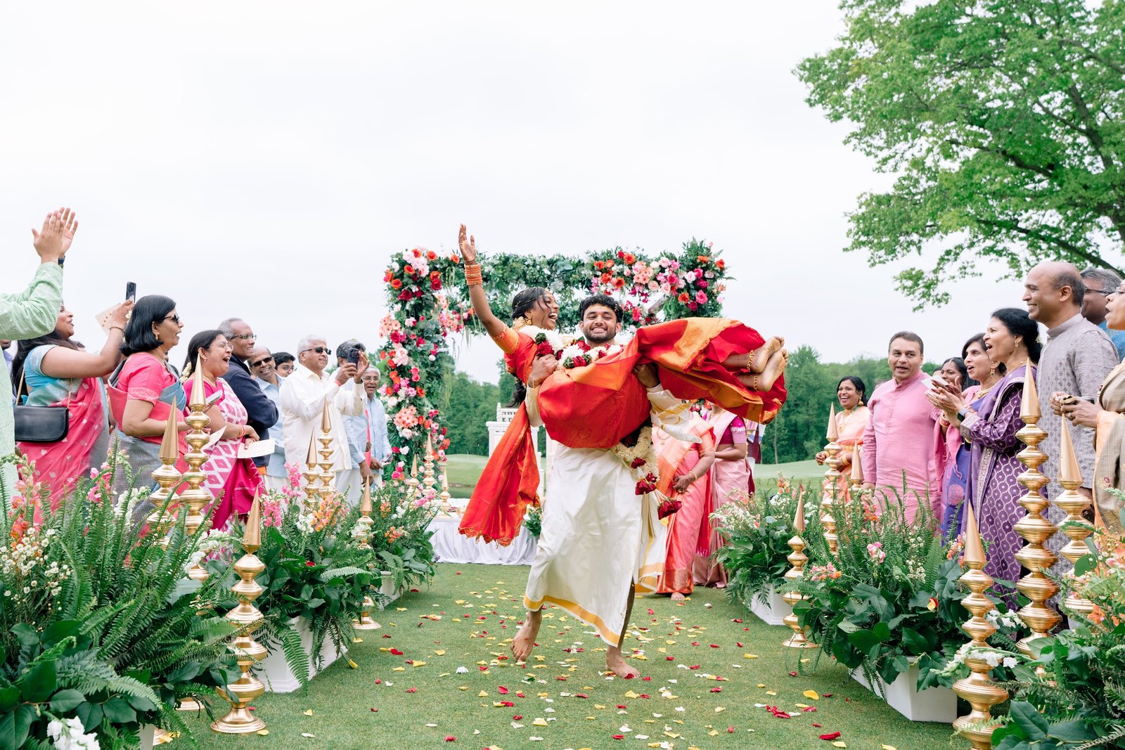 Groom lifted by family as guests cheer along a floral aisle