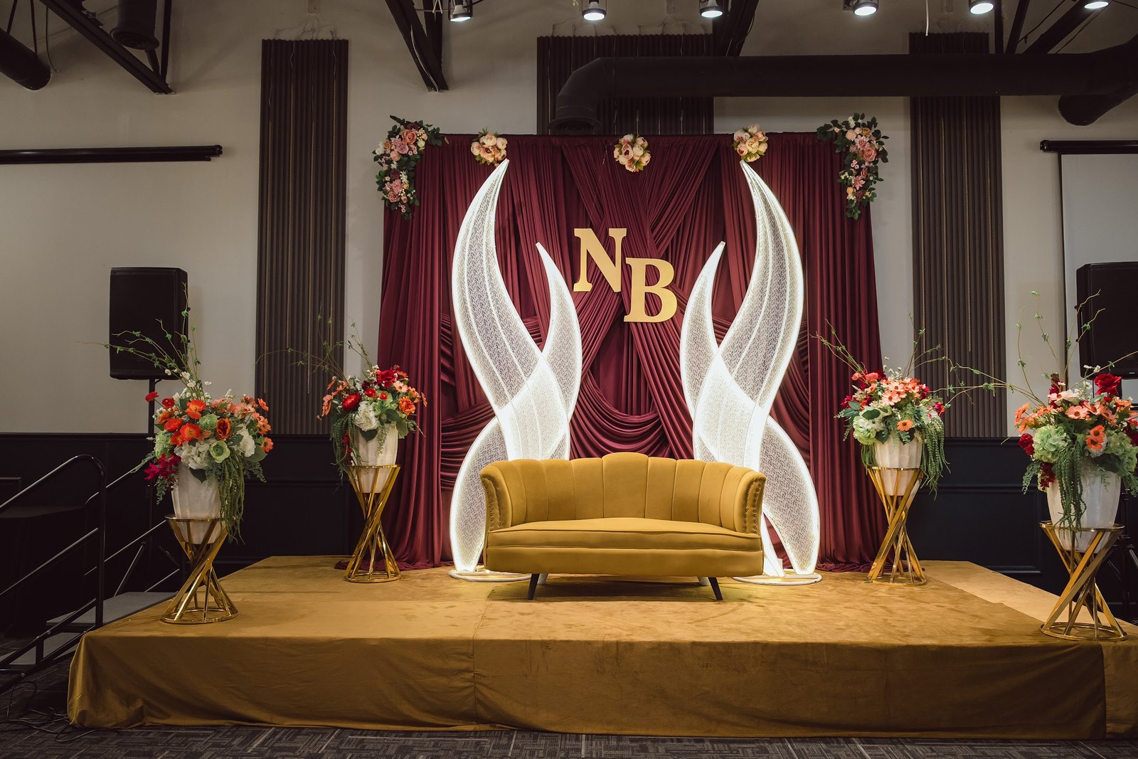 Maroon draped stage with lit white wings, a gold monogram and gold sofa