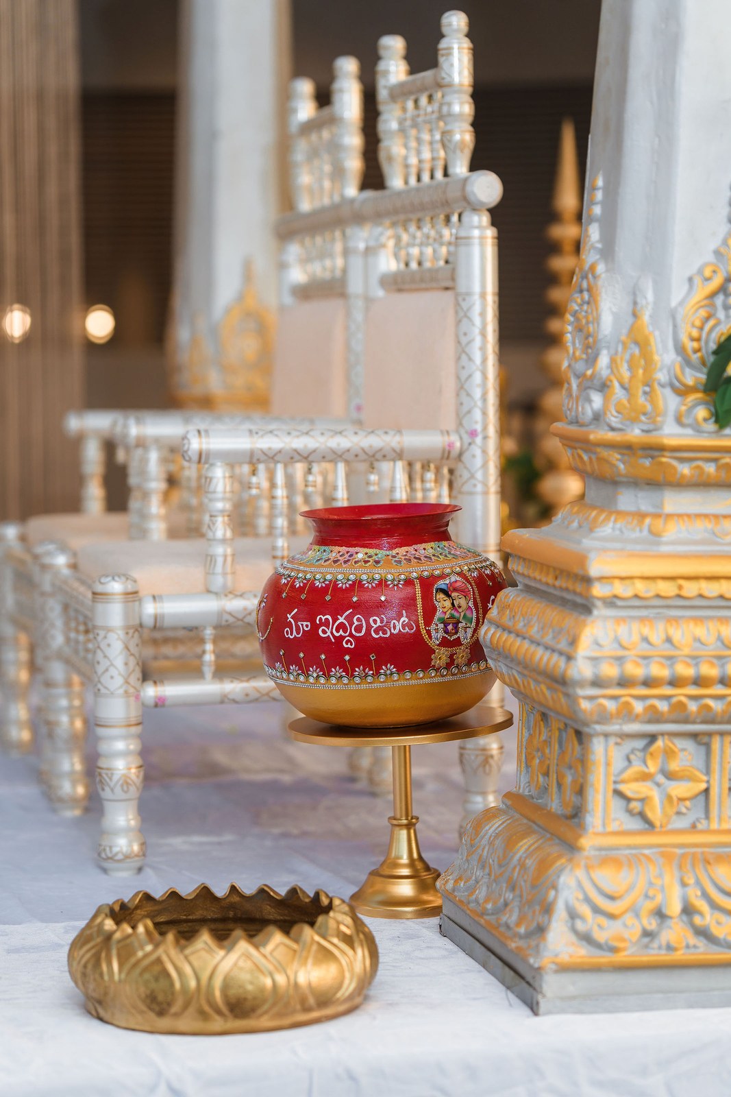 Hand-painted red kalash on a gold stand beside a gold lotus urli and carved mandap pillar