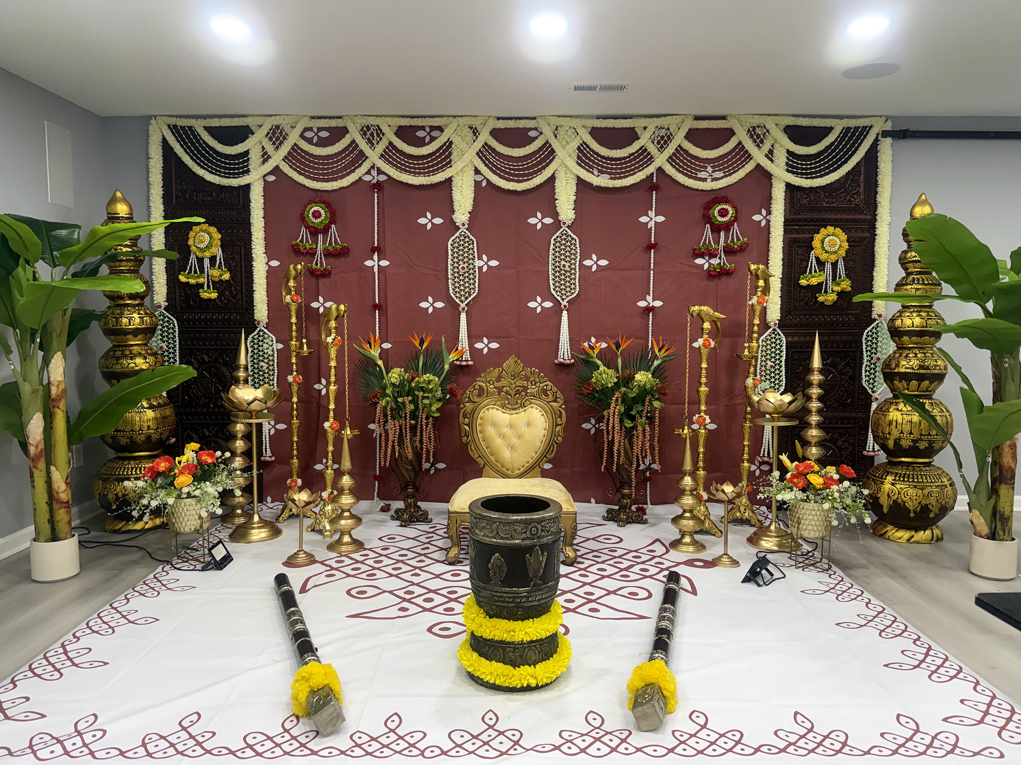 Traditional ceremony stage with a maroon backdrop, jasmine garlands, carved gold pillars, brass lamps and a marigold-wrapped mortar and pestles