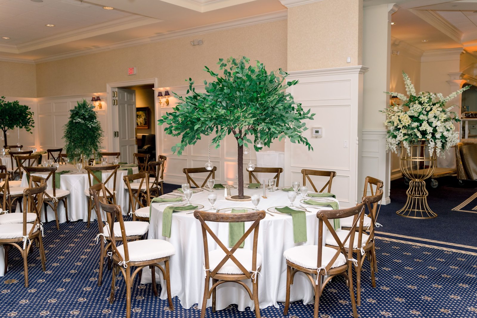 Reception tables with tall green tree centerpieces and cross-back chairs